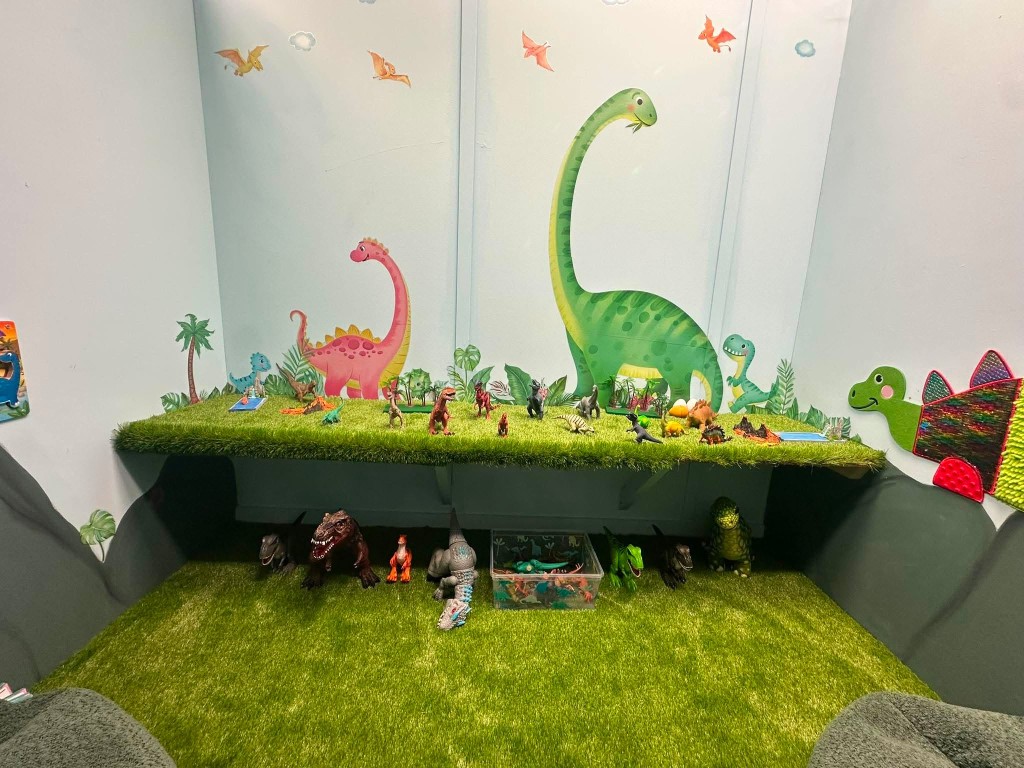 Dinosaur figurines on a grass shelf, with painted dinosaurs on the wall behind.