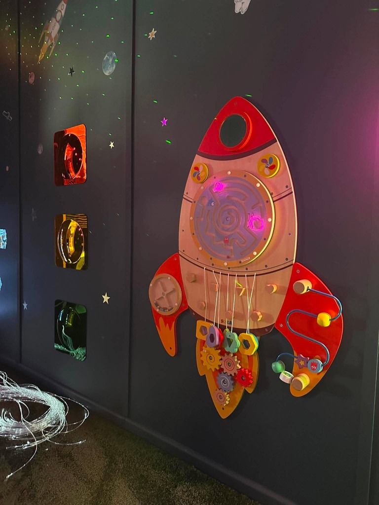 The space sensory room, with a blue bubble tube, a wooden rocket, fibre optic lights and star projections.