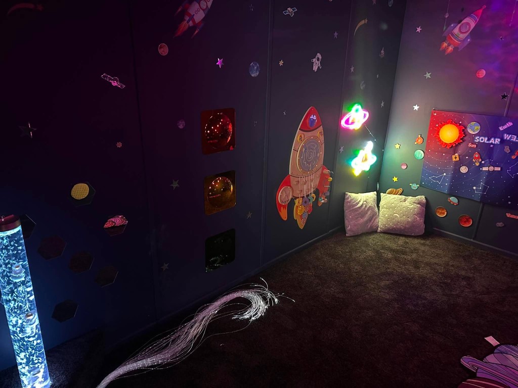 A wooden rocket on the space wall, with gears, a bead maze and fibre optic lights on the floor.