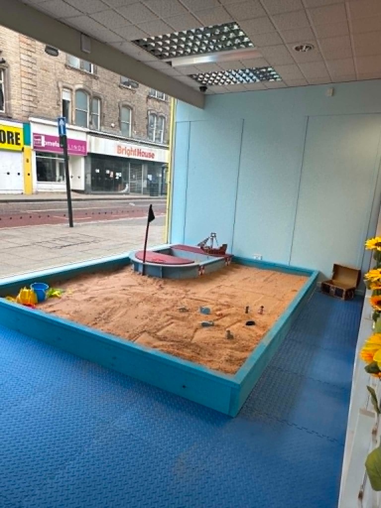 The sand pit with a toy boat and a wooden treasure chest, beside the high-street window.