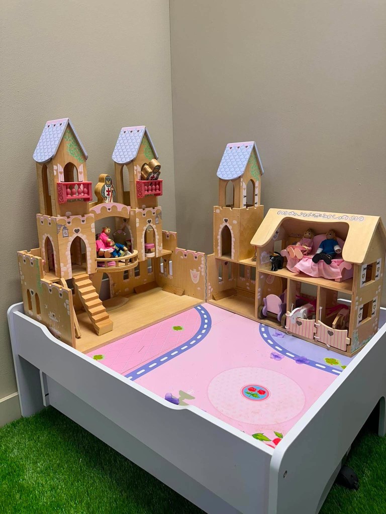 A wooden toy palace on a pink play table, with little figures in the towers and rooms.