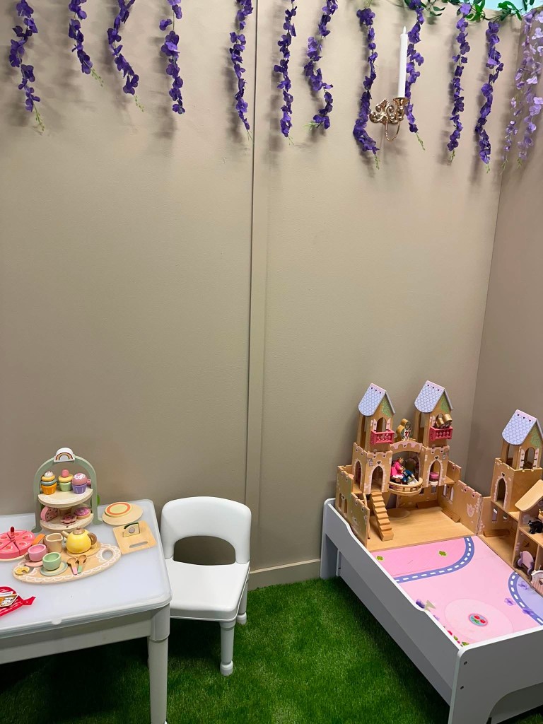 A wooden toy castle and a tea party table on the grass, with purple flowers on the wall.