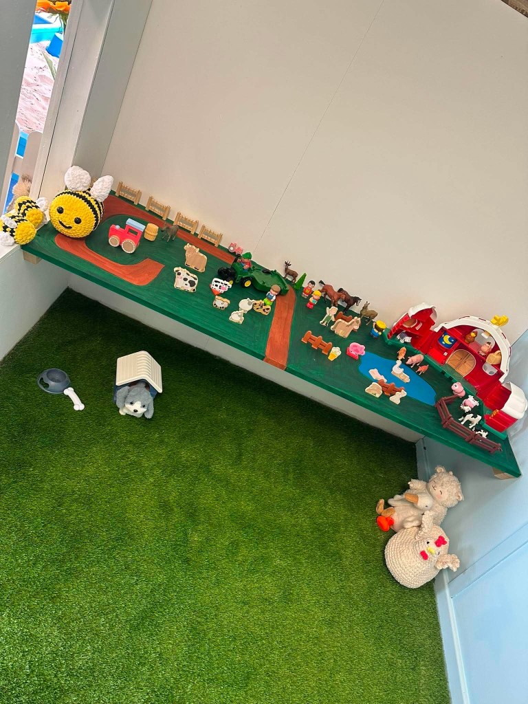 A grass farm shelf of animals, a red barn and crocheted bees, with a sheep and hen on the floor.