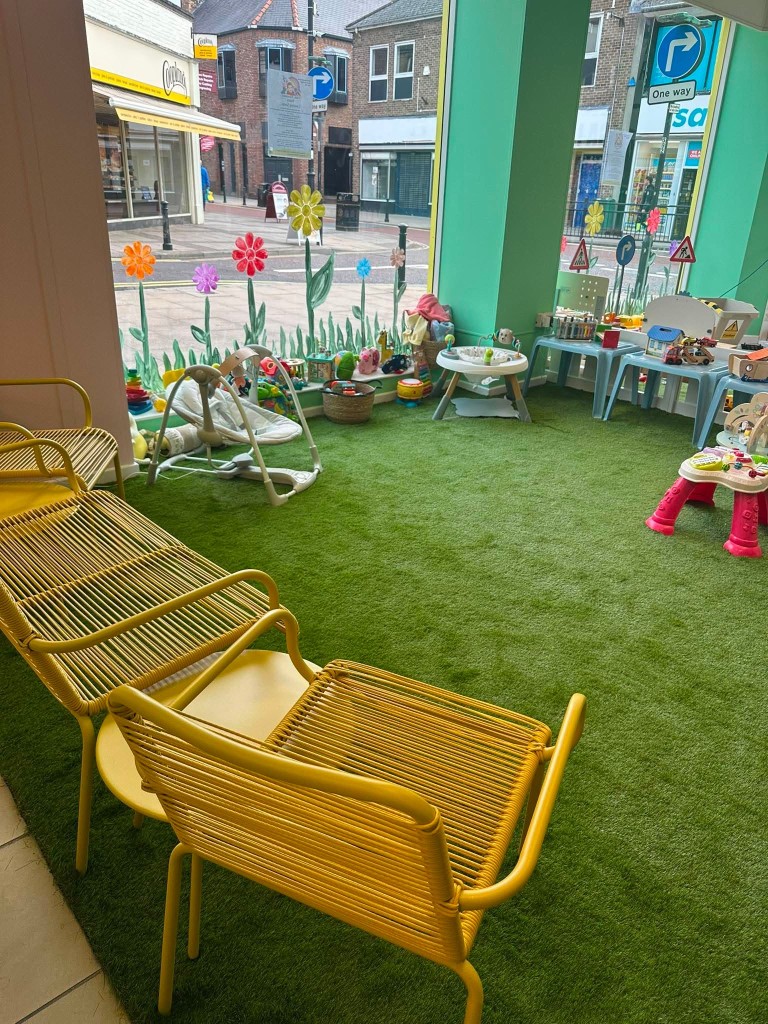 The grass play floor by the high-street windows, with yellow chairs for grown-ups and toys for little ones.