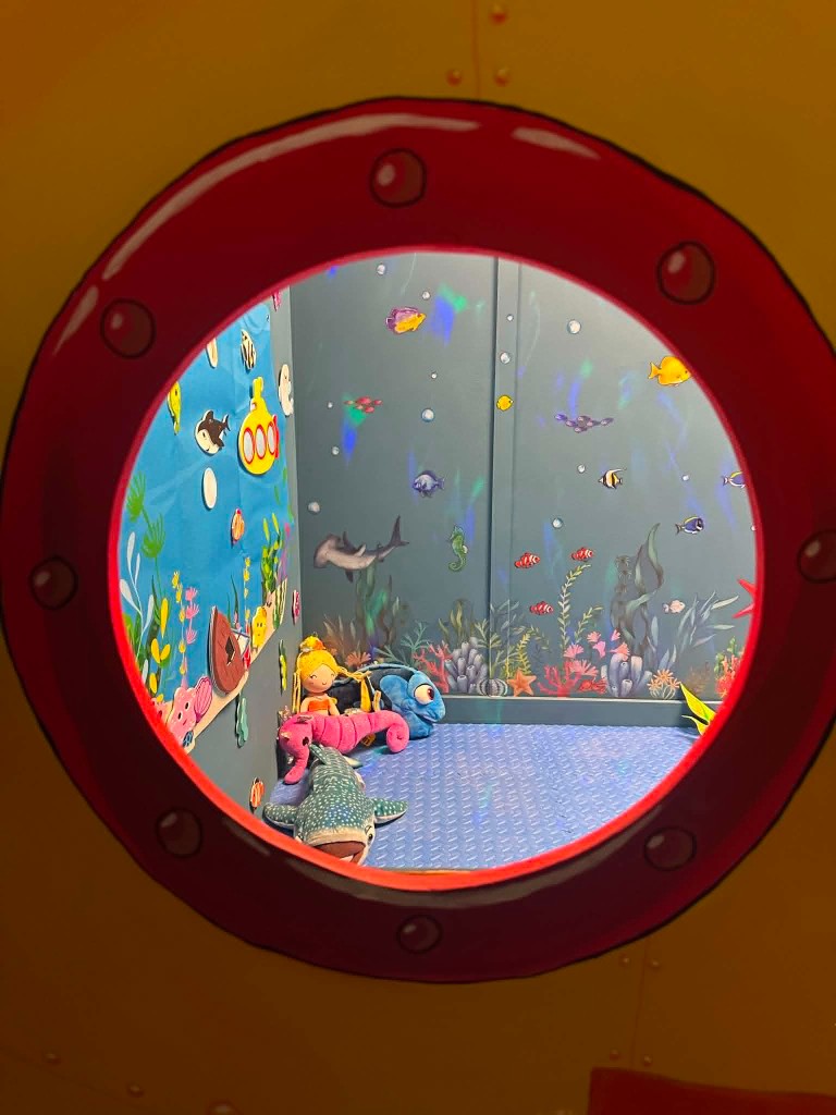 Looking through a red porthole into the underwater play room, with a painted reef and sea creature toys.