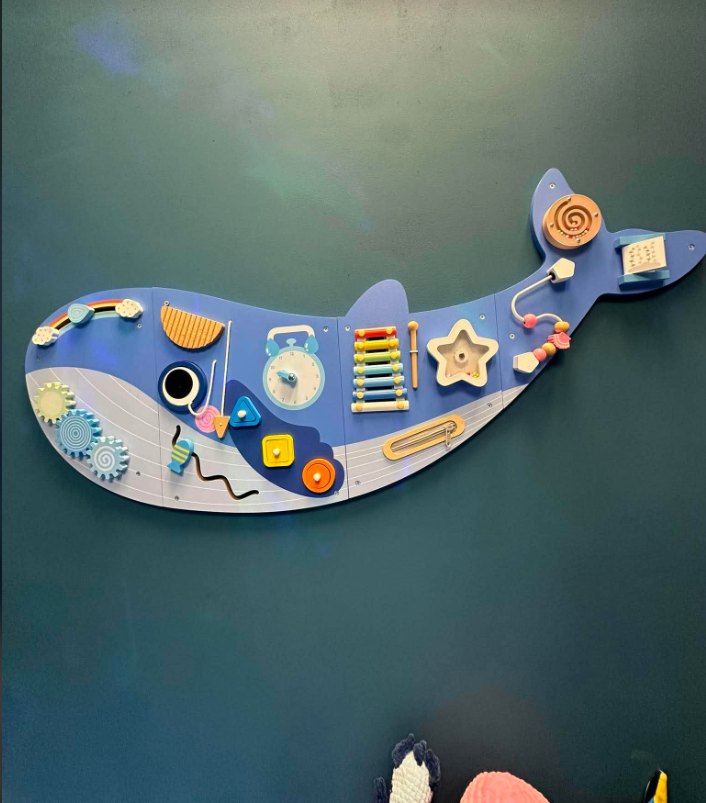 A blue whale activity board on the sea wall, with gears, a xylophone and bead mazes.